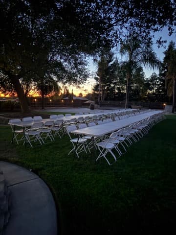 Long white tables and folding chairs arranged on a lawn at sunset.