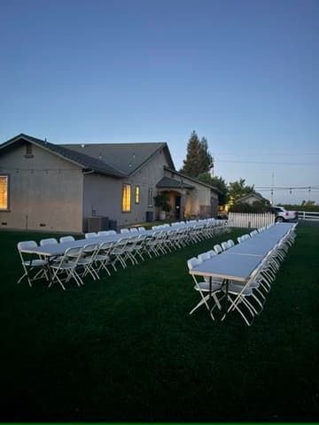 Three long rows of white folding tables and chairs arranged on a lawn at dusk.
