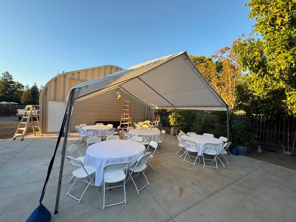 White canopy over round tables with white linens and folding chairs on a driveway.