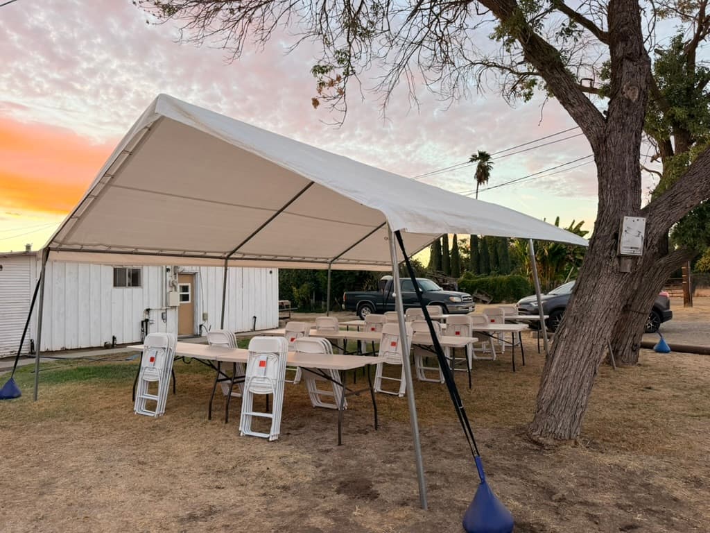 Wide white canopy with folding tables and stacked white chairs beneath a pink sunset sky.