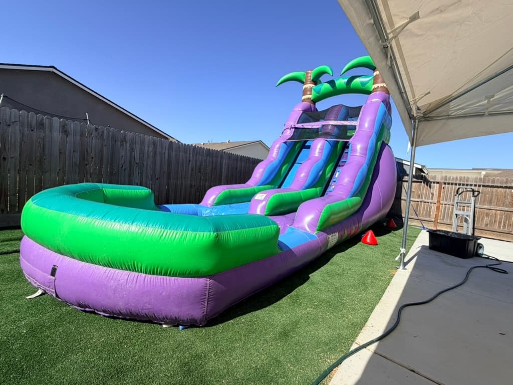 Purple, green, and blue palm-tree water slide on artificial turf beneath a clear sky.